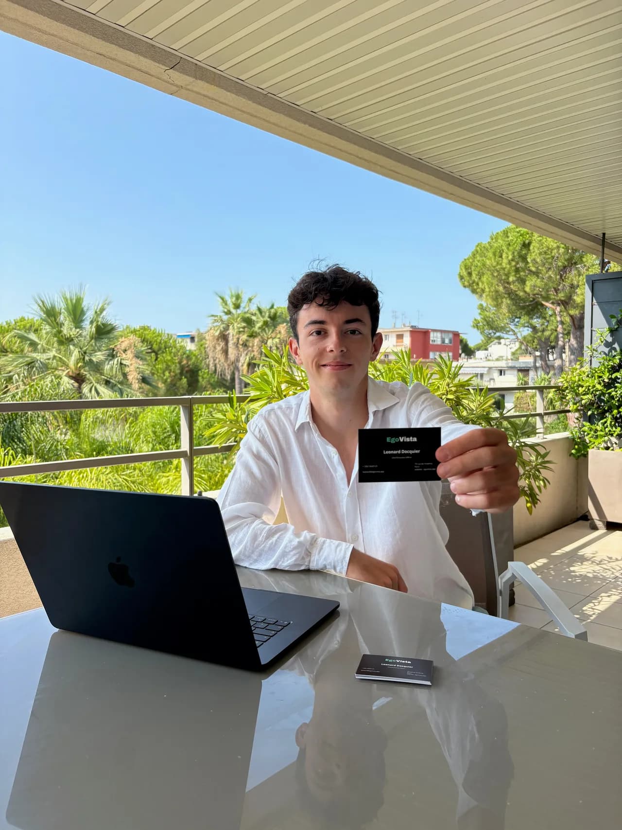 A young man in a white shirt seated at a glass table on a sunny terrace with palm trees behind him, an open laptop to his left, holding out a black business card toward the camera.